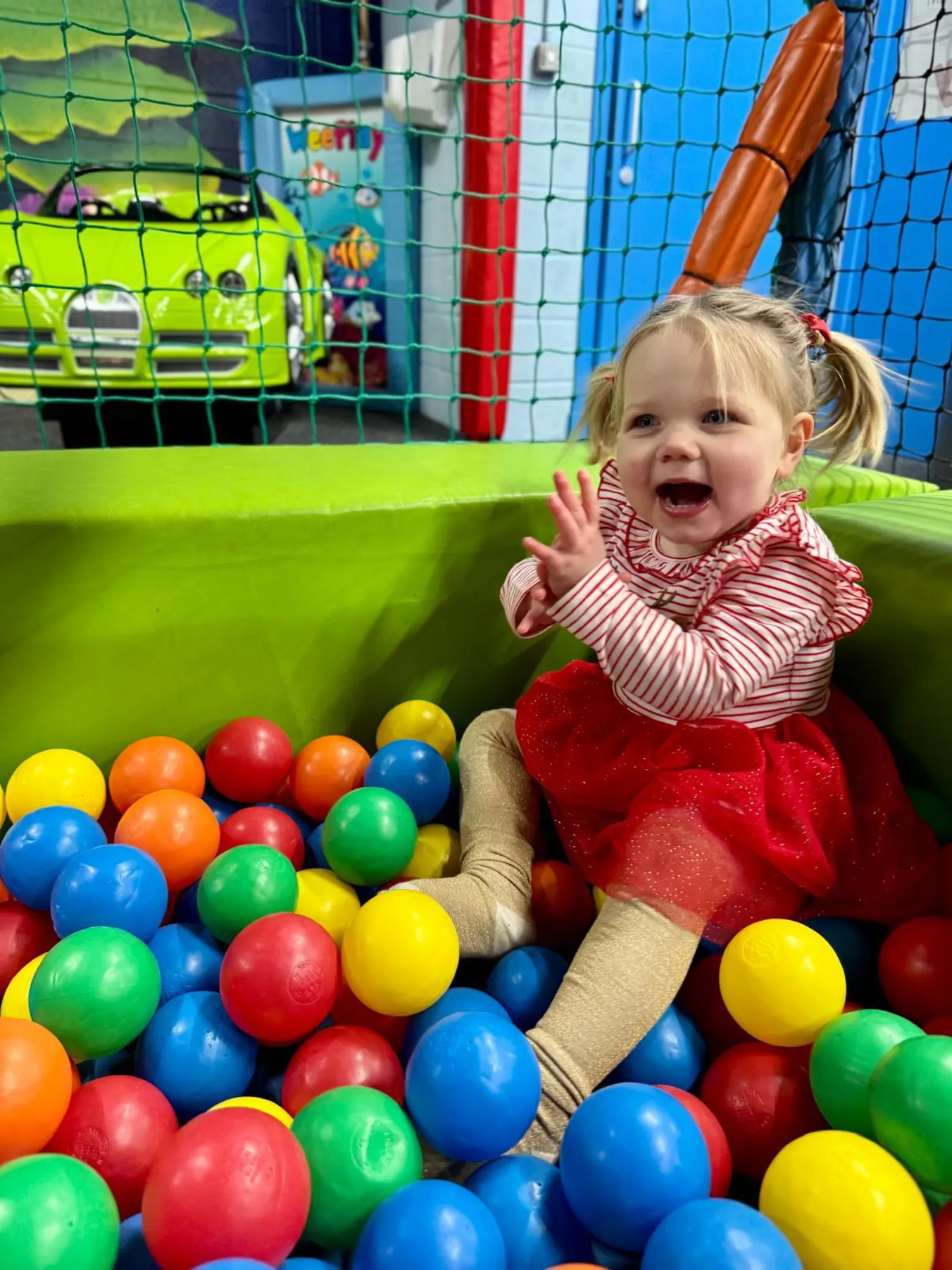 A delighted toddler in a red dress laughing in a ball pit full of bright balls